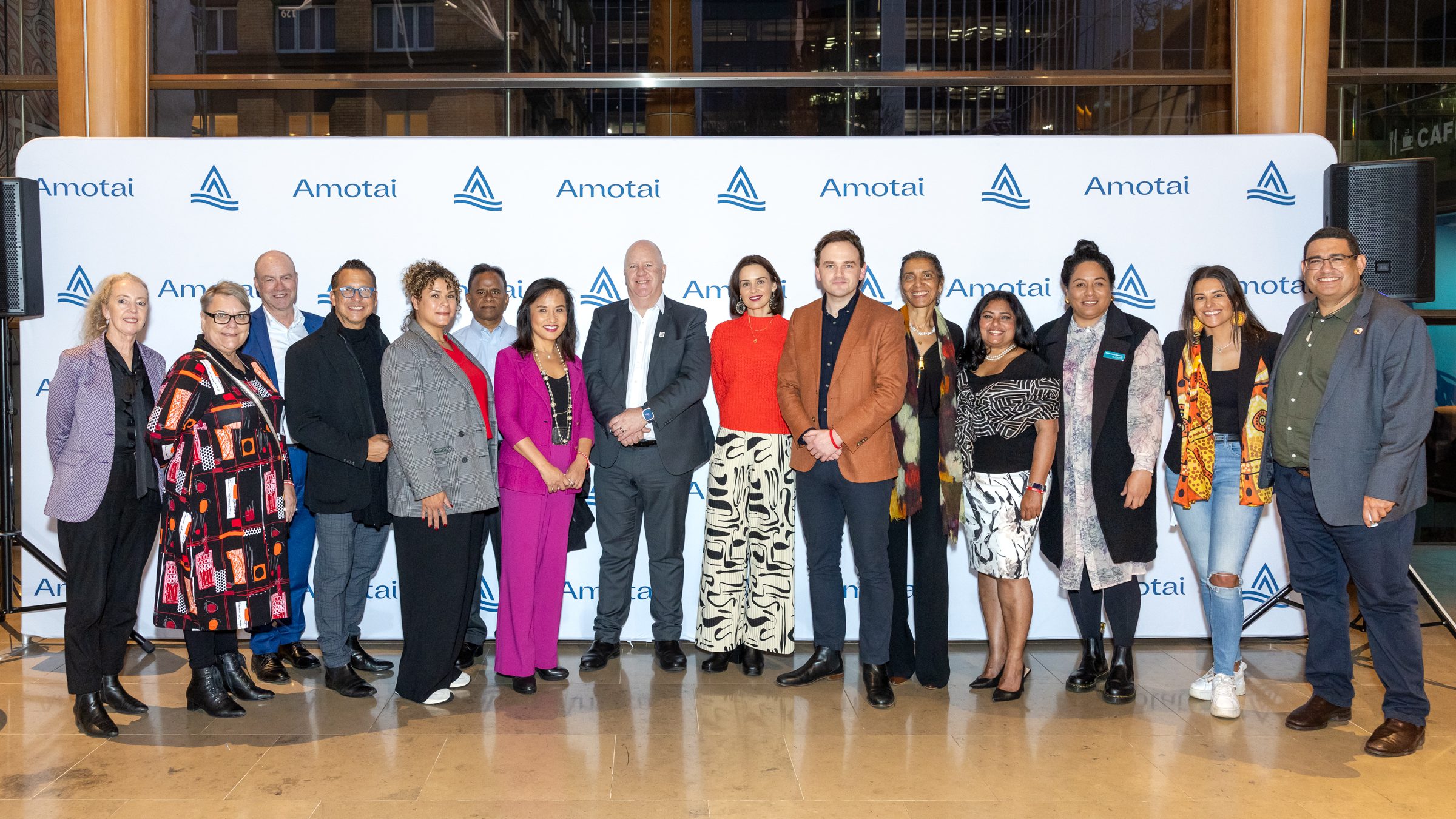 Group photo of men and women of a variety of races against a step and repeat banner with the words Amotai in blue against a white background. 